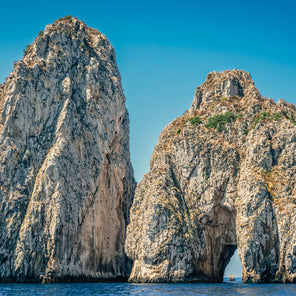 A photograph of two large rock structures in coastal Italy with boats sailing toward and through the overpass. 