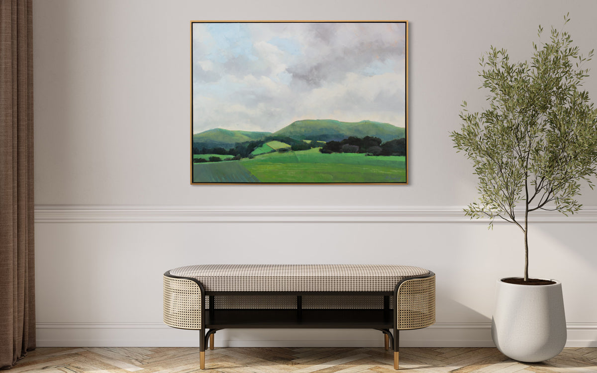 A light grey and shades of green landscape painting by Molly Doe Wensberg hangs in a light wood floater frame on a white wall above a modern rattan bench and a tall potted plant.