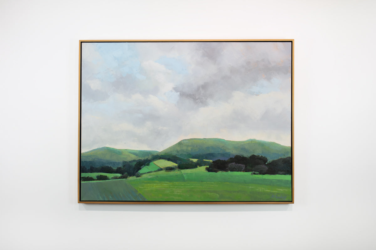 A light grey and shades of green landscape painting by Molly Doe Wensberg hangs in a light wood floater frame on a white wall.