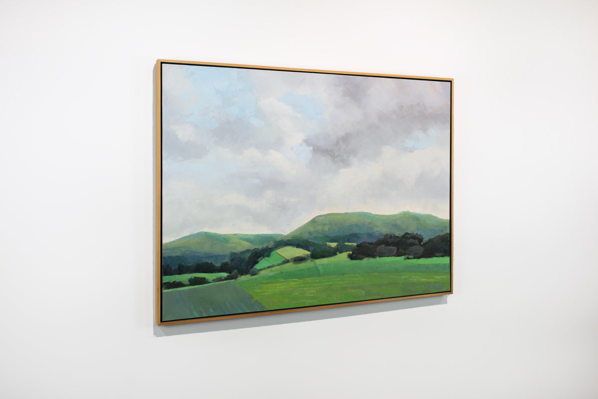 A side view of a light grey and shades of green landscape painting by Molly Doe Wensberg hangs in a light wood floater frame on a white wall.