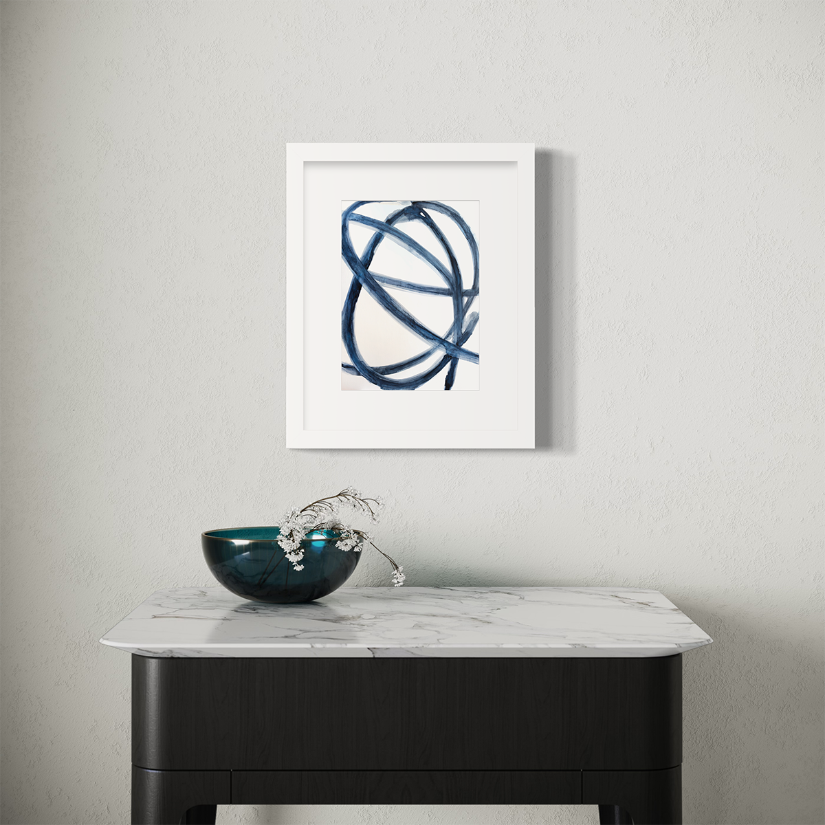 A blue and white abstract print in a white frame with a mat hangs on a white wall above a white marble table with a teal bowl with floral stems.
