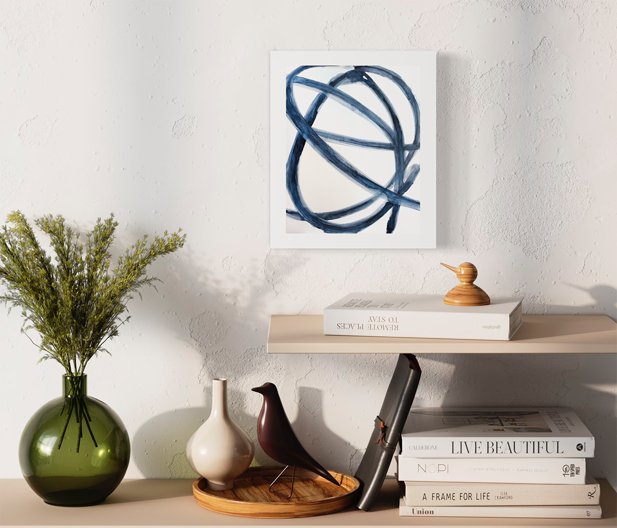 A blue and white abstract print in an unmatted white frame hangs on a white wall above two wood shelves with books, a tray with decor items and a green vase with greenery.