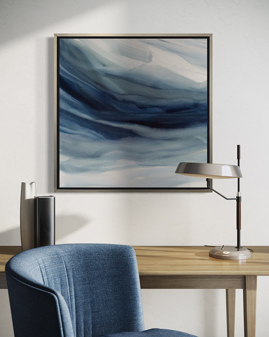 A blue, navy and white abstract print in a silver floater frame hangs on a white wall above a maple table with a silver lamp and decorative vases. A blue chair is is front.
