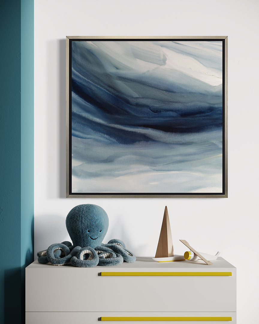 A blue, navy and white abstract print in a silver floater frame hangs on a white wall above a white childs chest of draws with a stuffed blue octopus and some wooden toys.