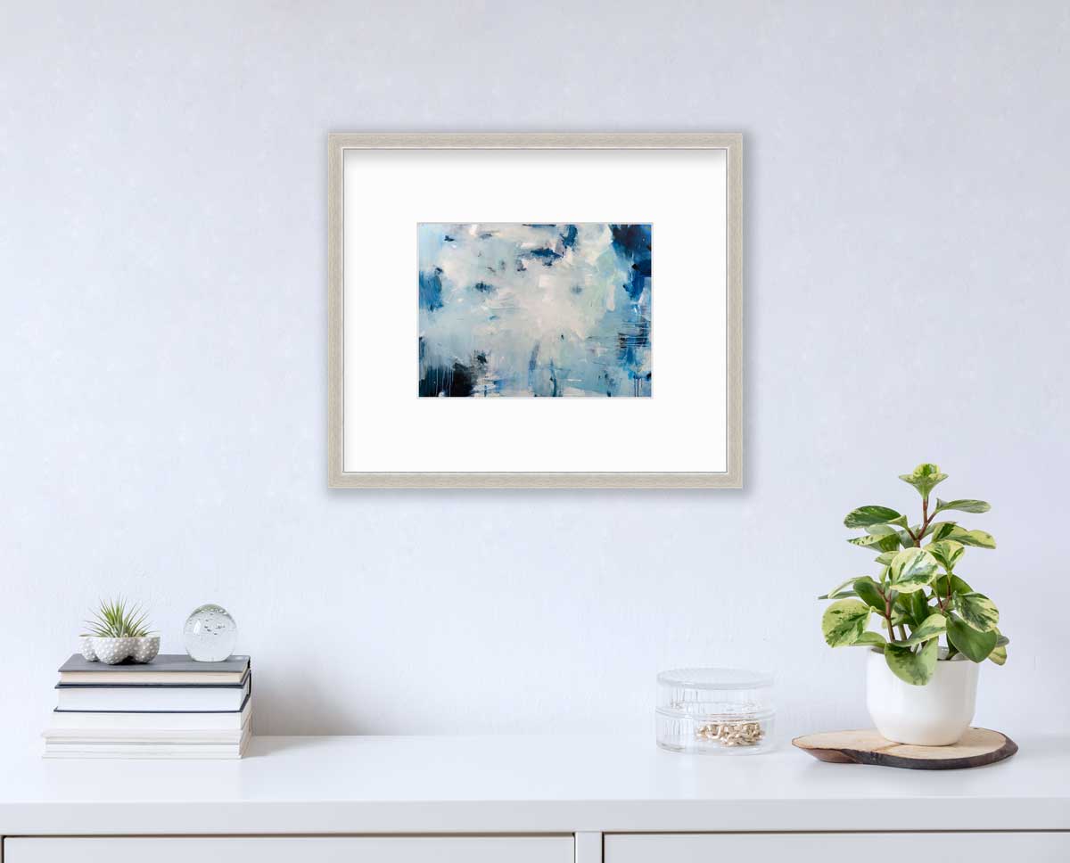 A blue and white abstract print by Kelly Rossetti in a silver frame with a mat hangs on a white wall above a white surface with a stack of books, a round clear container with gold accessories and a potted plant.