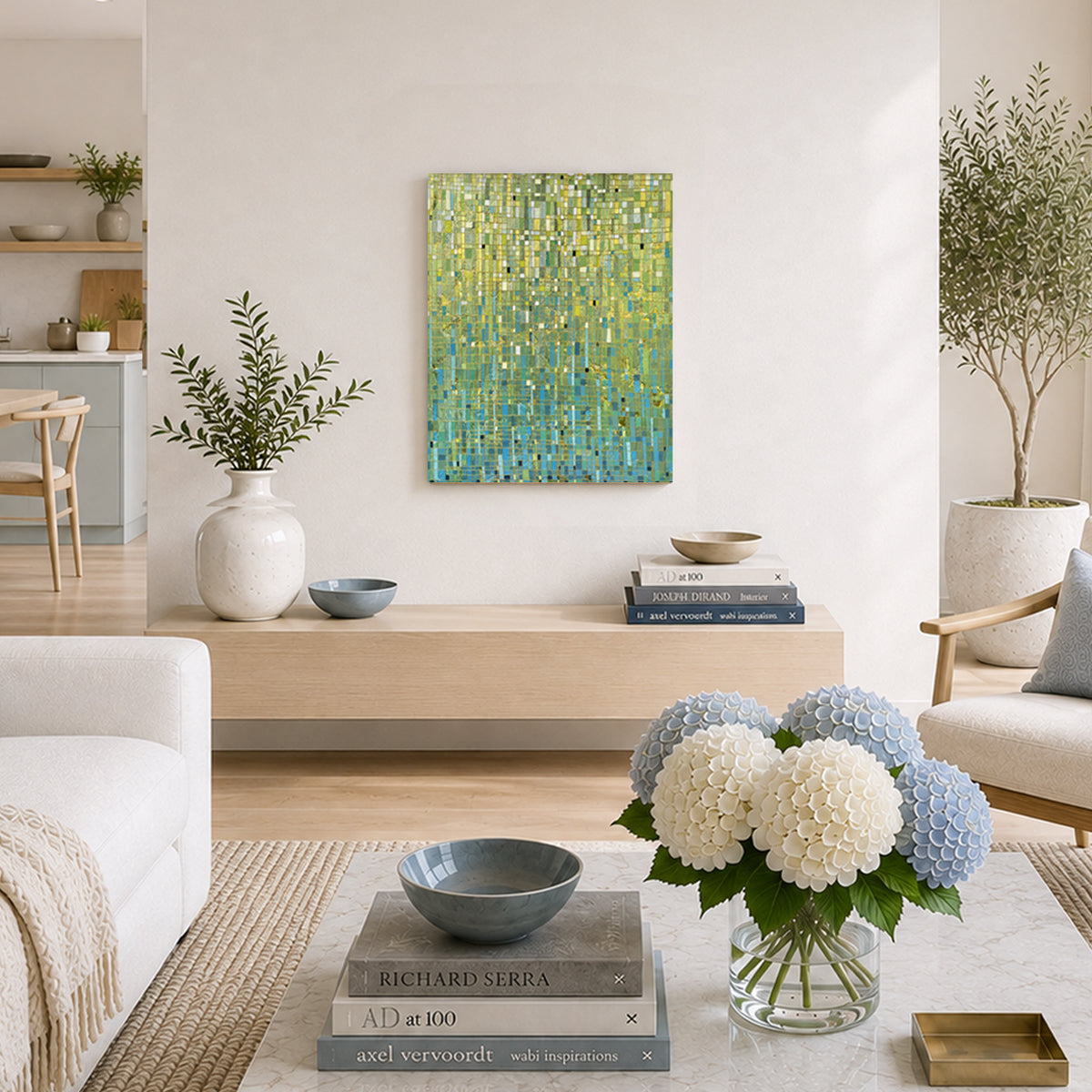 An abstract painting with a textured green and blue brushstroke pattern hangs on a light beige wall in a modern, sunlit living room. The room features a light wood console table, a white sofa, a marble coffee table with blue and white hydrangeas, and potted plants.