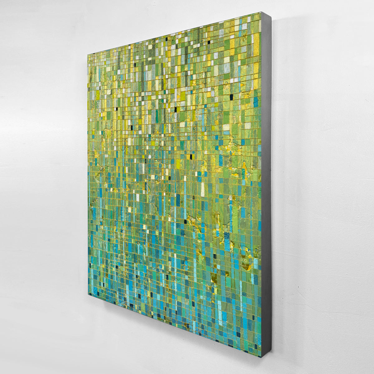 An abstract painting with an abstract geometric pattern in shades of green, yellow, and blue, shown hanging on a light gray wall.