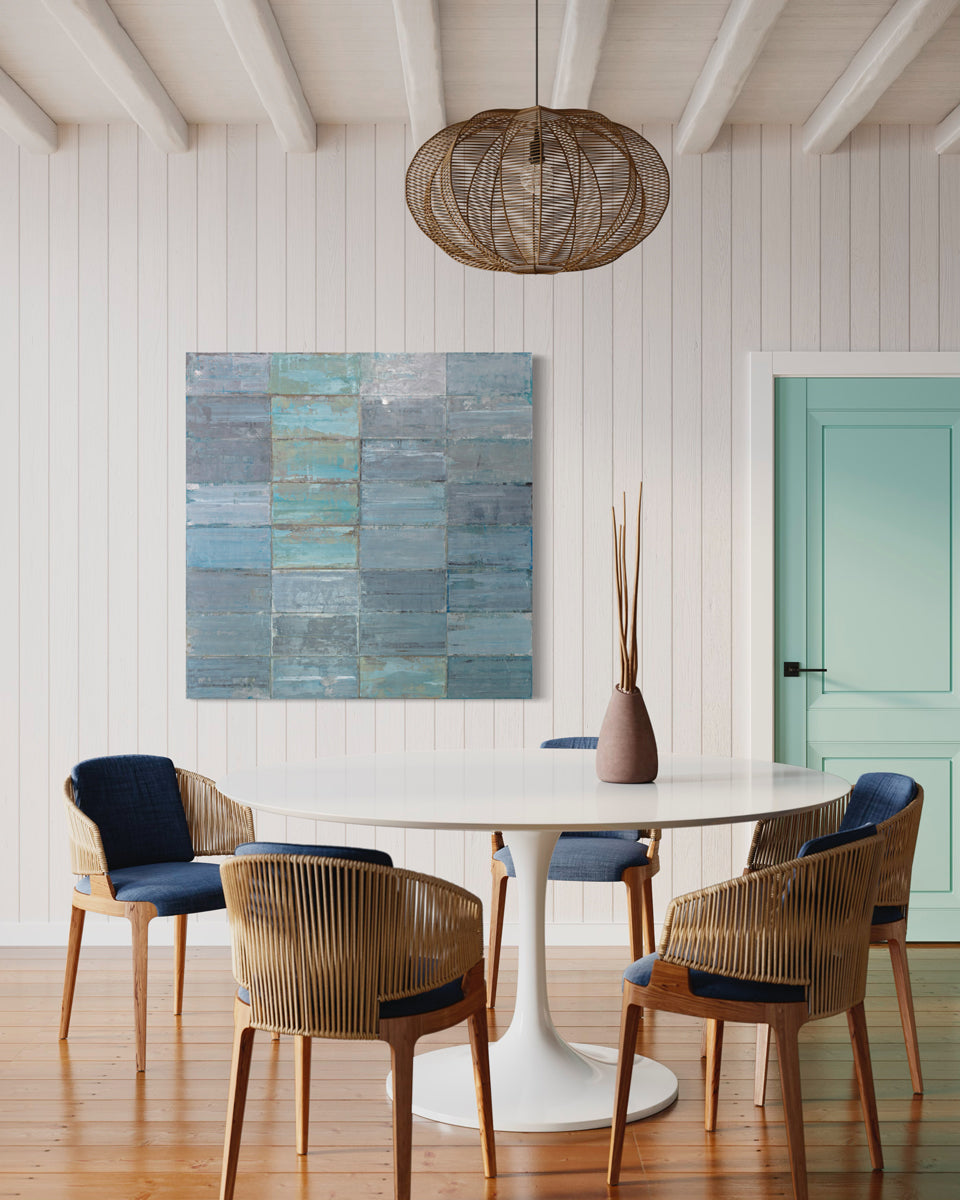 A blue and grey abstract geometric painting hangs on a white paneled wall above a midcentury white dining room table with blue wicker chairs and beneath a coastal woven hanging light fixture. 