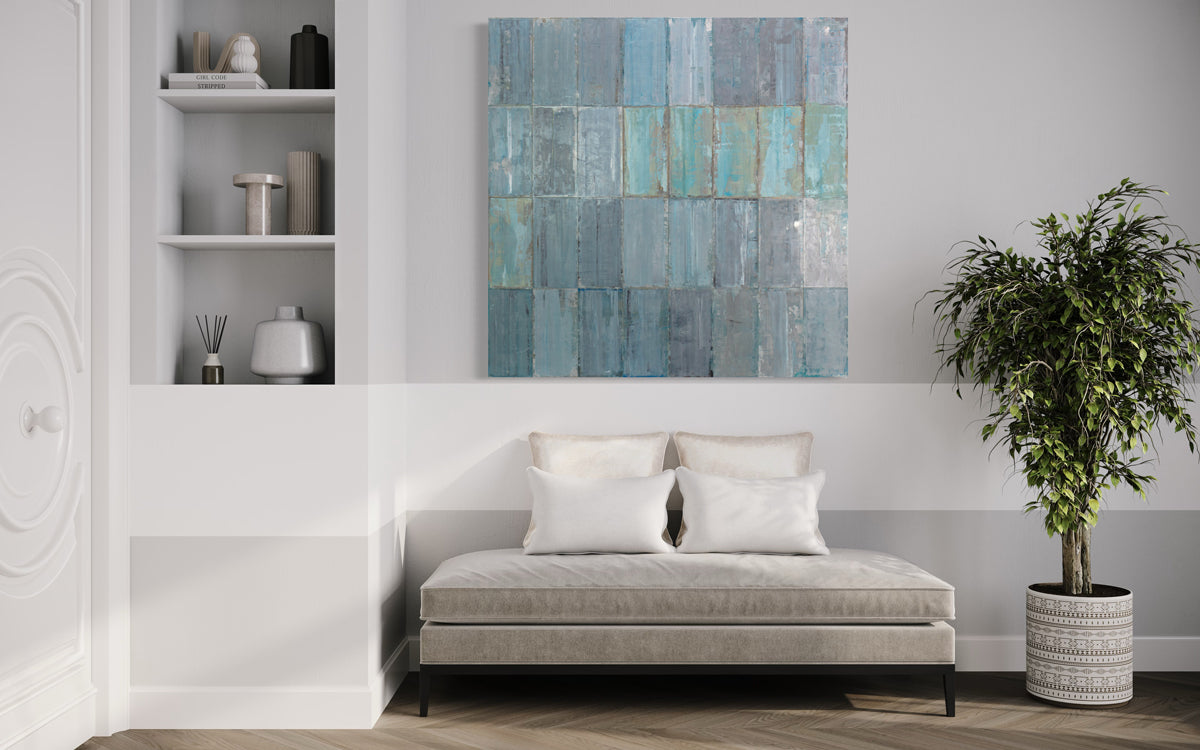 A blue and grey abstract geometric painting hangs on a white wall above a modern grey chaise and floor plant and next to built in shelving with modern decor. 