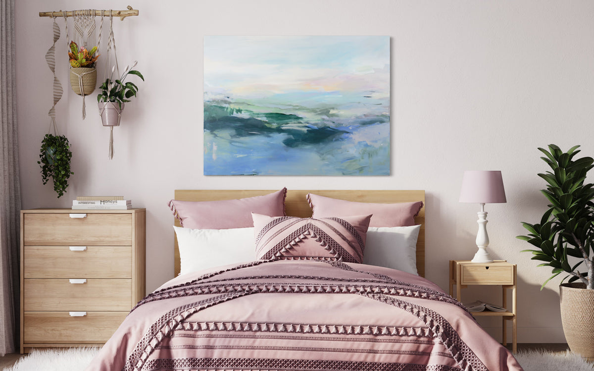A blue, green, and pink abstract painting hangs on a white wall above a bed with wooden frame and pink sheets, wooden dresser and end table with a white and pink lamp, and a large potted floor plant. 