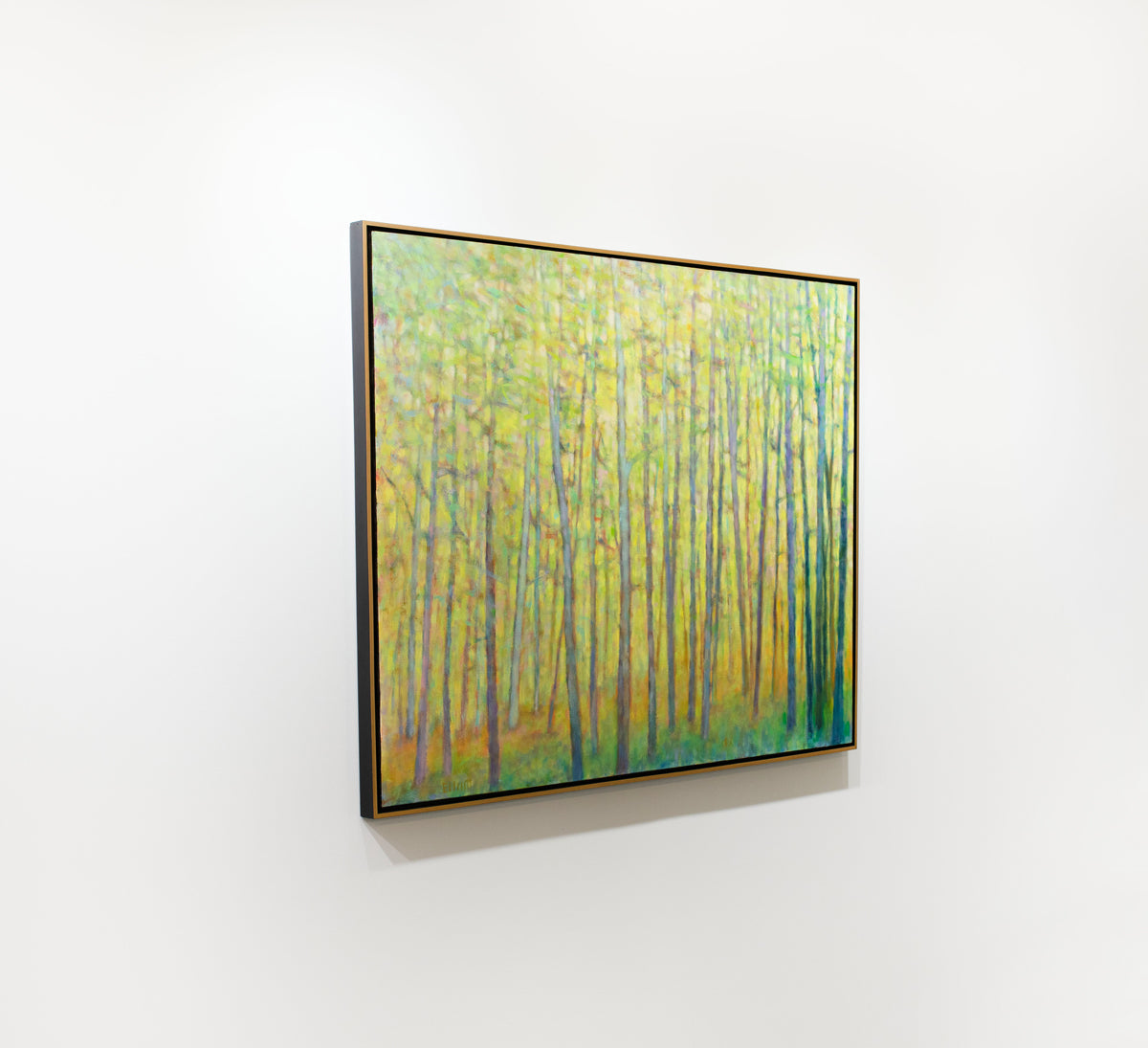 A yellow abstracted forest trees painting framed in a thin gold floater frame hangs on a white wall.