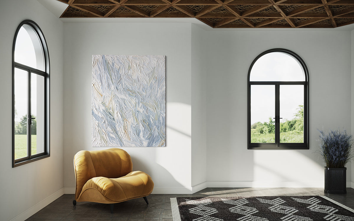 An abstract heavily textured painting with blue, grey, white, stone, teal, and yellow ochre hangs on a white wall in natural light by Teodora Guererra. This painting hangs over a yellow ochre velvet modern chair. To the left and right are windows and in front is a carpet in a black and white pattern. To the extreme right is a plant of lavender in a black ribbed pot.