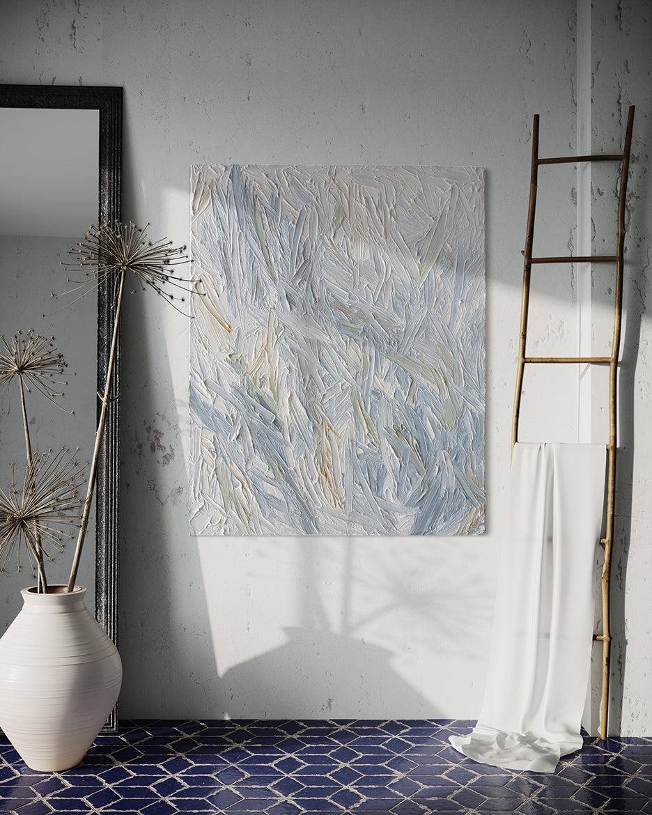 An abstract heavily textured painting with blue, grey, white, stone, teal, and yellow ochre paint hanging on a white stucco wall by Teodora Guererra. To the right is a natural wood decorative ladder with a white fabric draped over it. to the left is a wall mirror with a dark ridged frame leaning on the wall and a large white ceramic pot with decorative sticks in front. The floor below is a dark blue tile in a geometric pattern with grey grout.