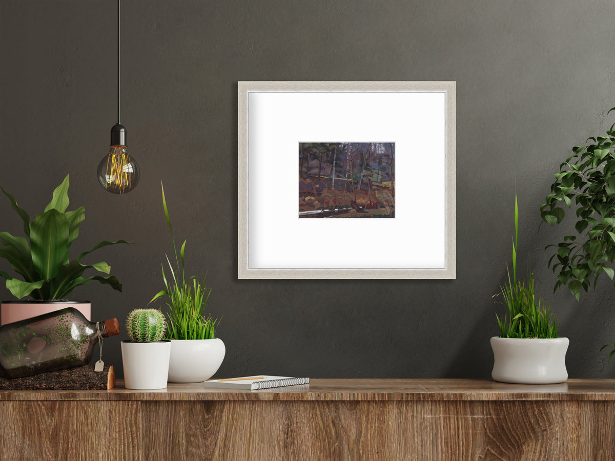 A brown, grey and green abstract landscape print in a silver frame with a mat hangs on a dark grey wall above a wood surface with several potted plants, a notebook and a decorative bottle.