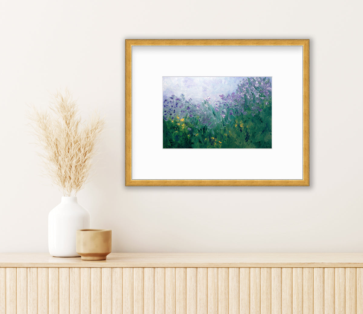 A blue, purple and green abstract print in a gold frame with a mat hangs on a white wall above a wood surface with a small beige cup and a white vase with wheat stems.