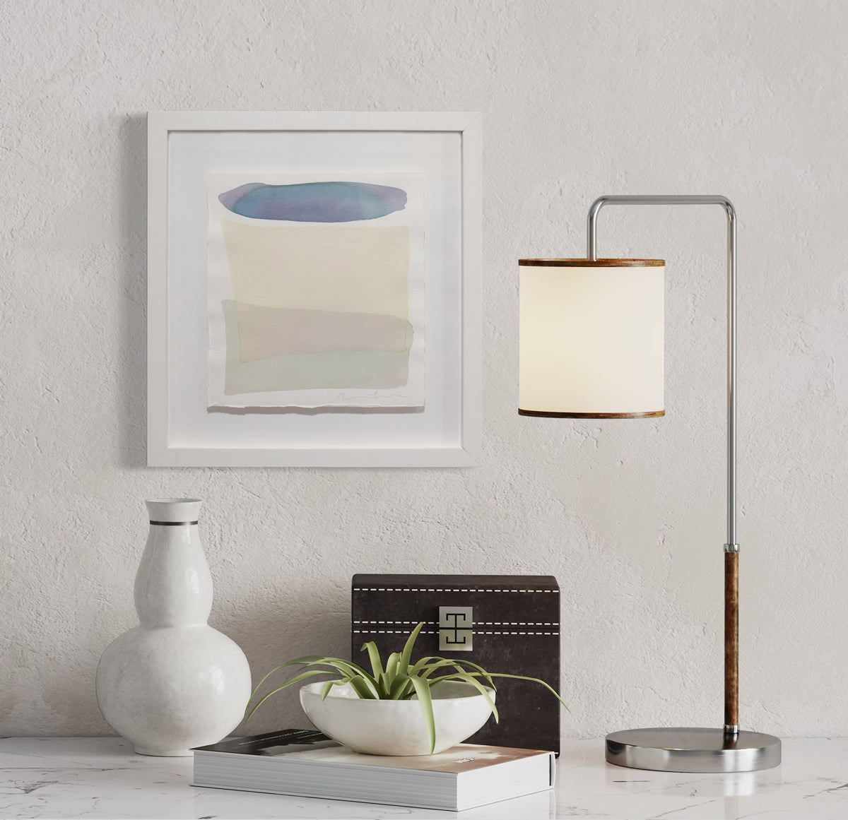 A teal blue with purple, pale yellow and light sea foam green shapes overlap in this painting by Nealy Hauschildt is float mounted in a white frame hanging above a white marble surface with a small lamp, a white vase, a black box, a book and a greenery filled small bowl.