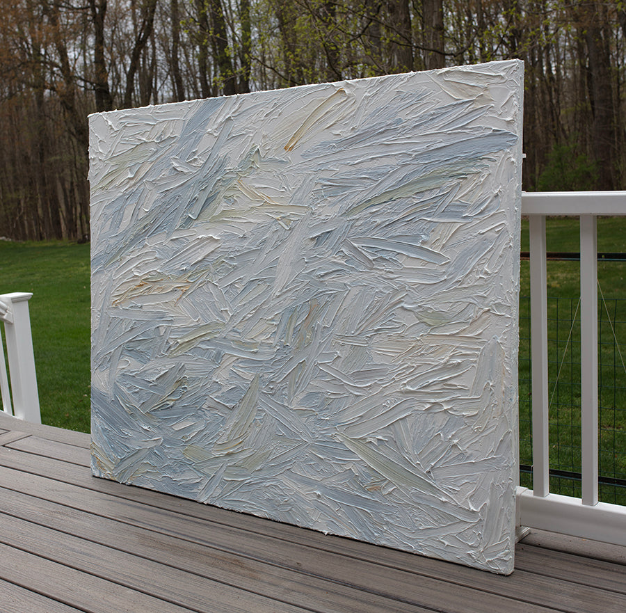 An abstract painting with thickly textured blueish, orangish and greenish beige brushstrokes is seen at an angle on the artists deck outside.