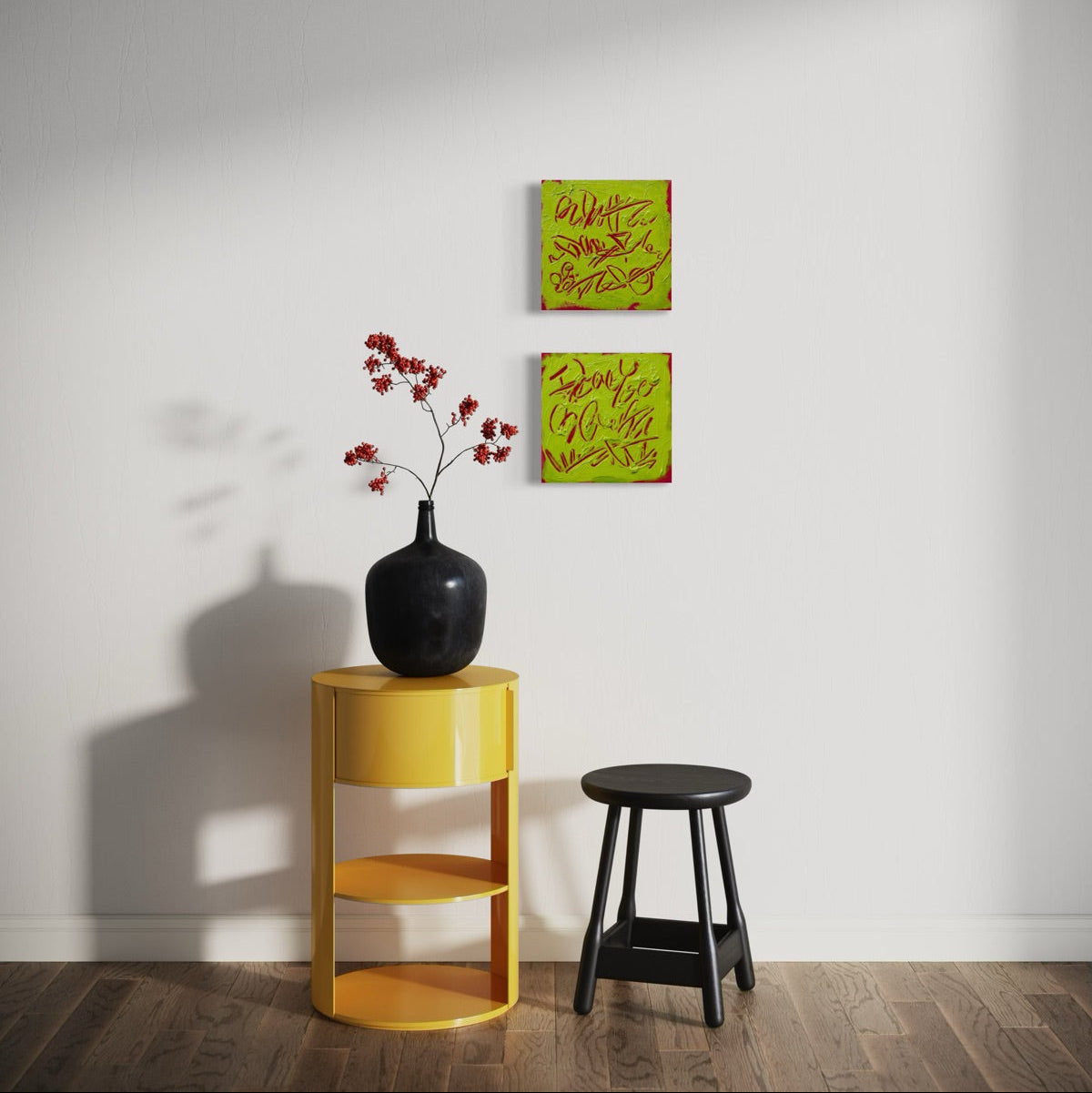 Two square abstract text-based paintings with textured chartreuse over hot raspberry pink  graffiti-style lettering, hang on a white wall above a yellow side table with a black vase of red flowers and a black stool.