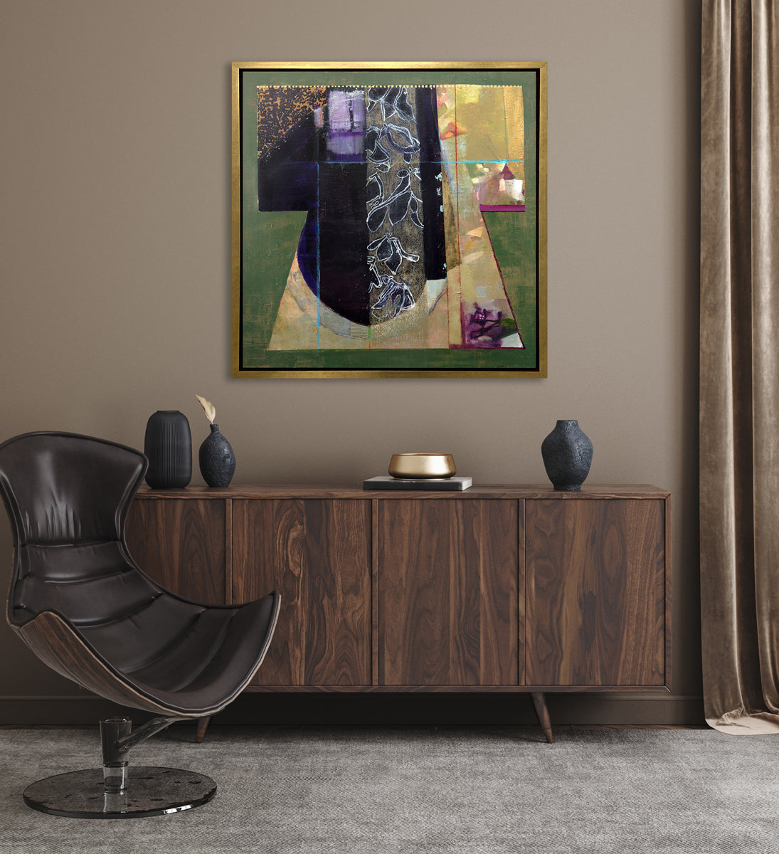 A green, gold and black abstract print in a gold floater frame hangs on a beige wall above a dark wood credenza with black and gold decorative accents and next to a deep brown leather modern chair.