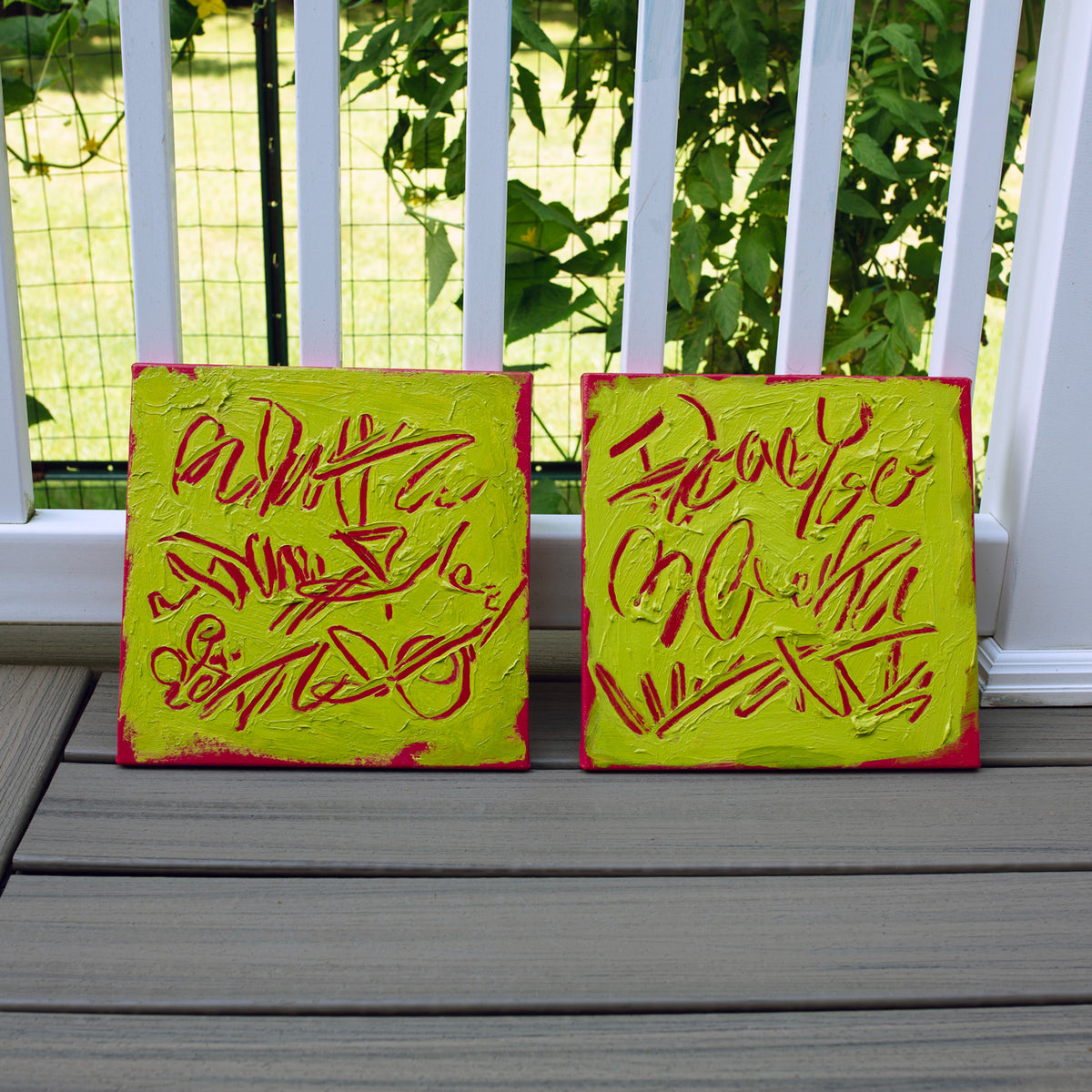 Two square abstract text-based paintings with textured chartreuse over hot raspberry pink  graffiti-style lettering, displayed side-by-side on a gray wooden deck in front of a white railing with green foliage in the background.