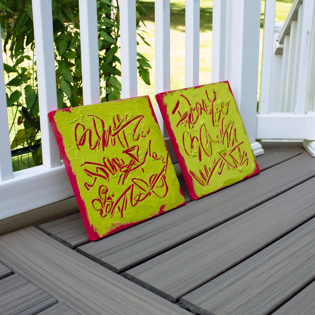 Two square abstract text-based paintings with textured chartreuse over hot raspberry pink  graffiti-style lettering, displayed side-by-side leaning on a gray composite deck railing outdoors. 