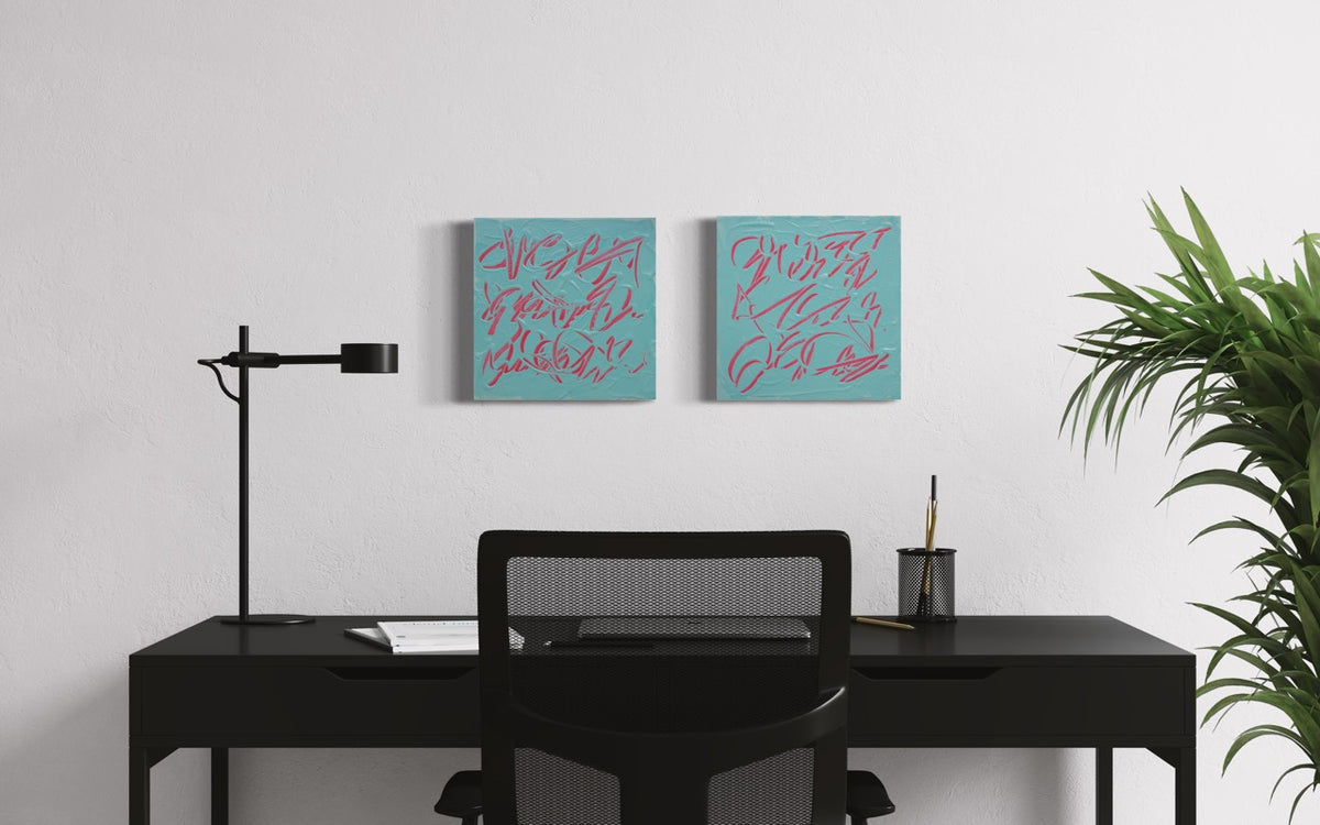 Two square abstract text based paintings with pink carved writing on a teal background, displayed side-by-side on a white wall above a black desk setup with a chair, lamp, and plant.
