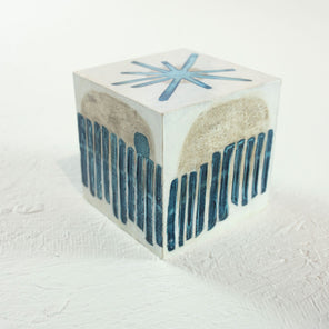 A white cube painted with blue and beige geometric shapes rests on a white surface. 