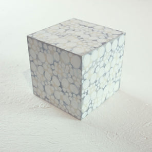 A wooden cube painted with overlapping white, beige, and grey circles rests on a white surface. 