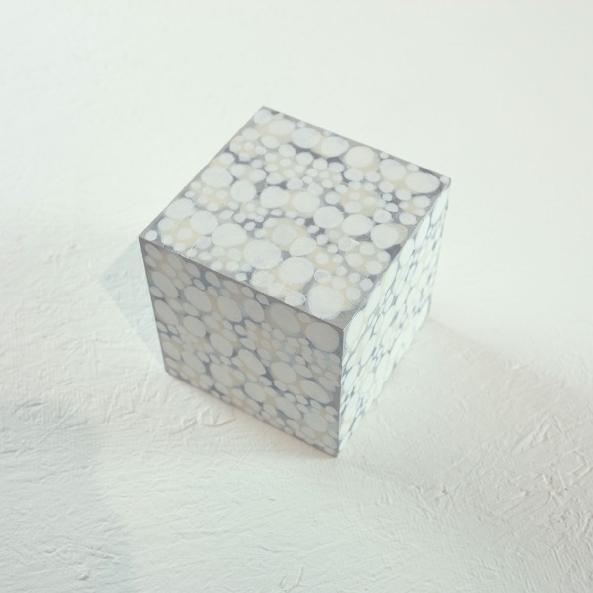 A wooden cube painted with overlapping white, beige, and grey circles rests on a white surface.
