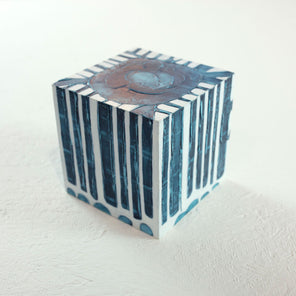 A white cube with a blue circle on the top and textured, geometric rectangles that extend over the sides of the cube rests on a white surface. 