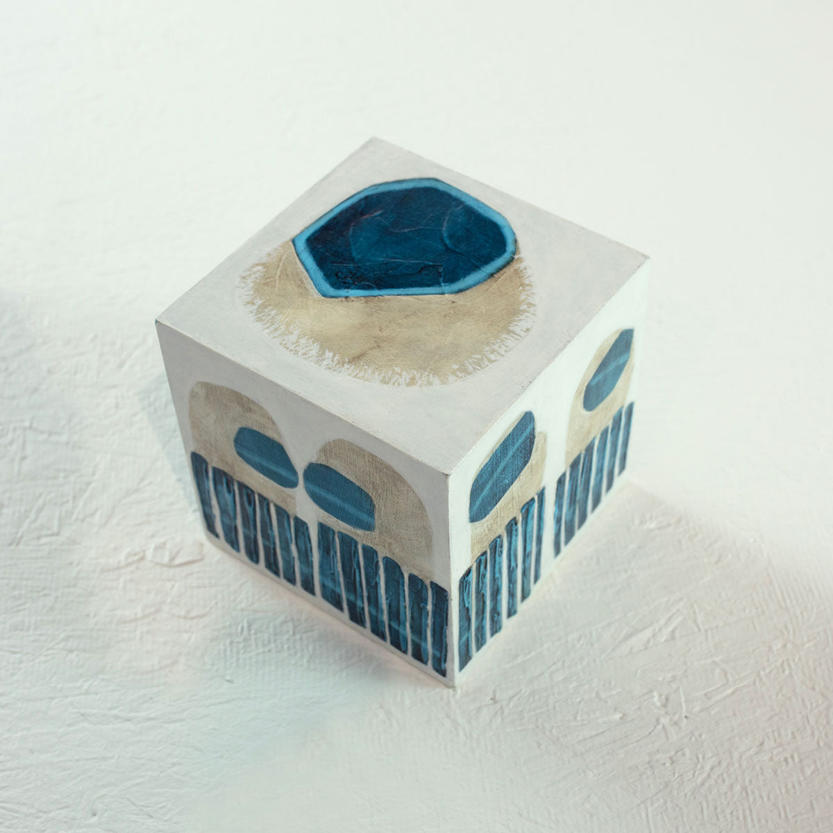 A white wooden cube with overlapping painted blue and beige shapes rests on a white surface.