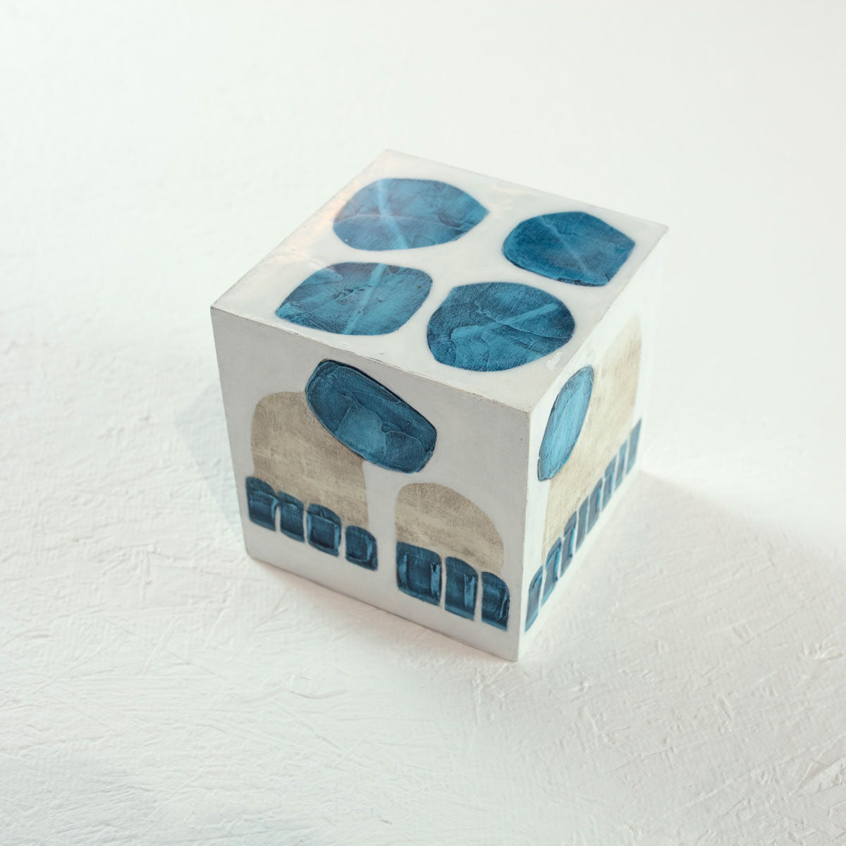 A white wooden cube with overlapping painted blue and beige shapes rests on a white surface.