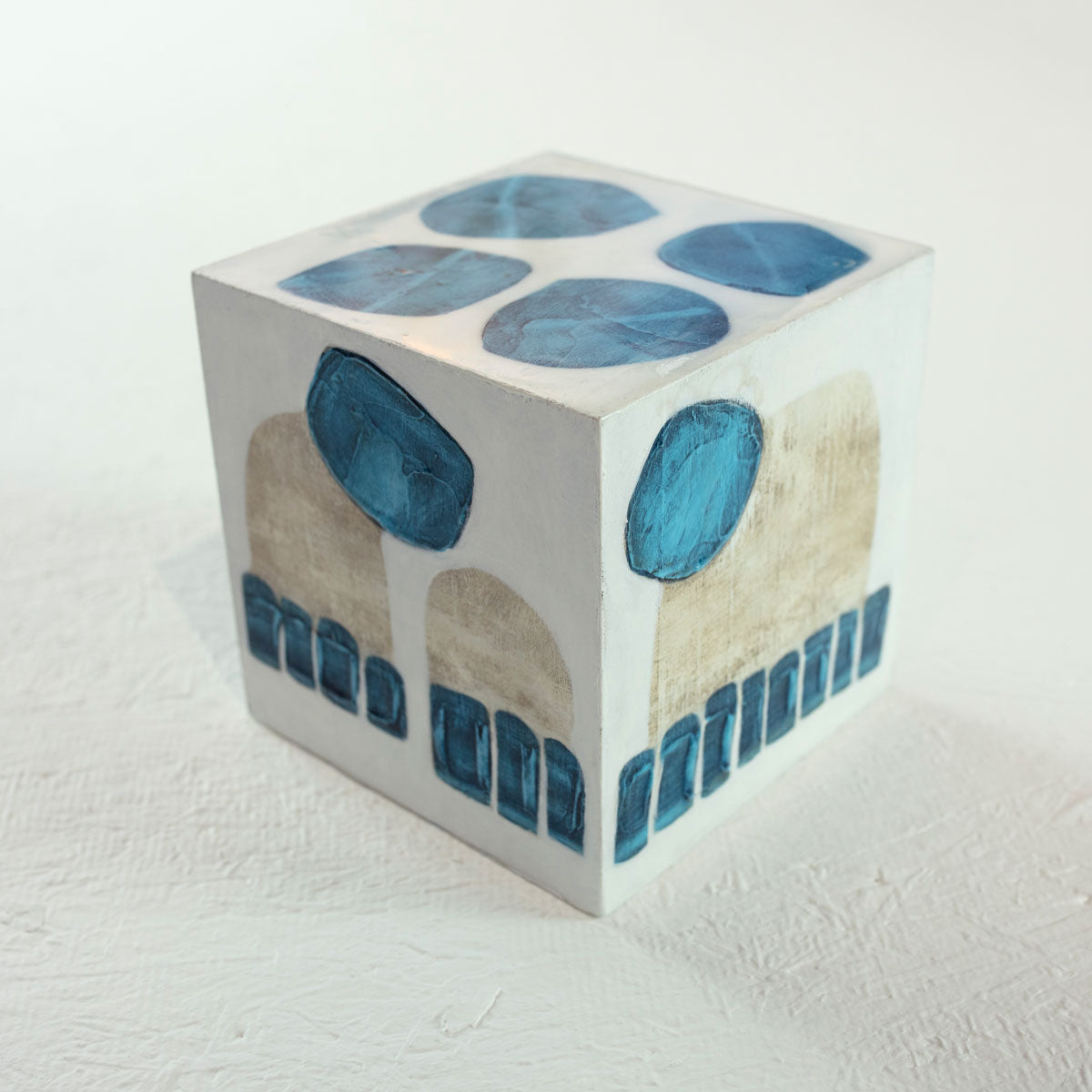 A white wooden cube with overlapping painted blue and beige shapes rests on a white surface.