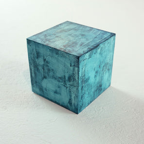 A painted textured turquoise cube rests on a white surface. 