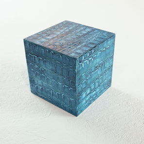A painted textured teal blue cube rests on a white surface.