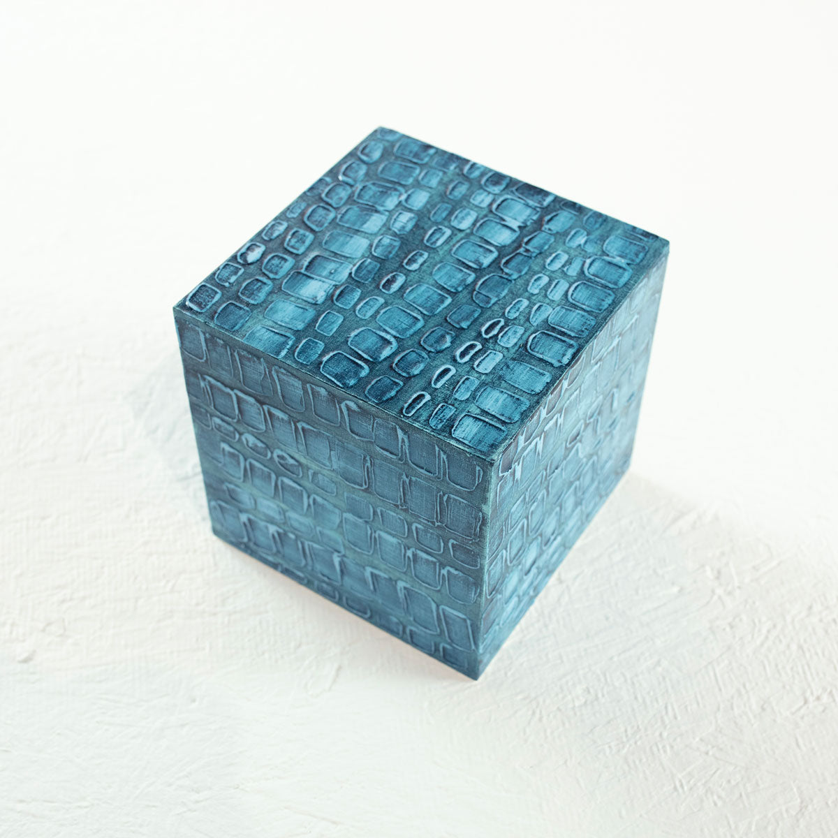 A painted textured teal blue cube rests on a white surface.