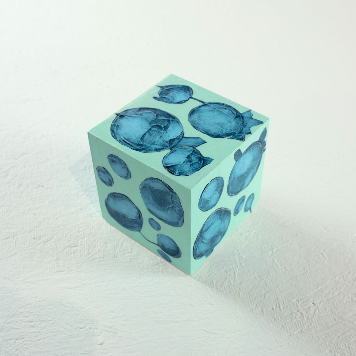A cube painted turquoise with imperfect blue circles rests on a white surface.