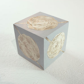 A grey cube with golden beige floral shapes painted on each face of the cube rests on a white surface. 