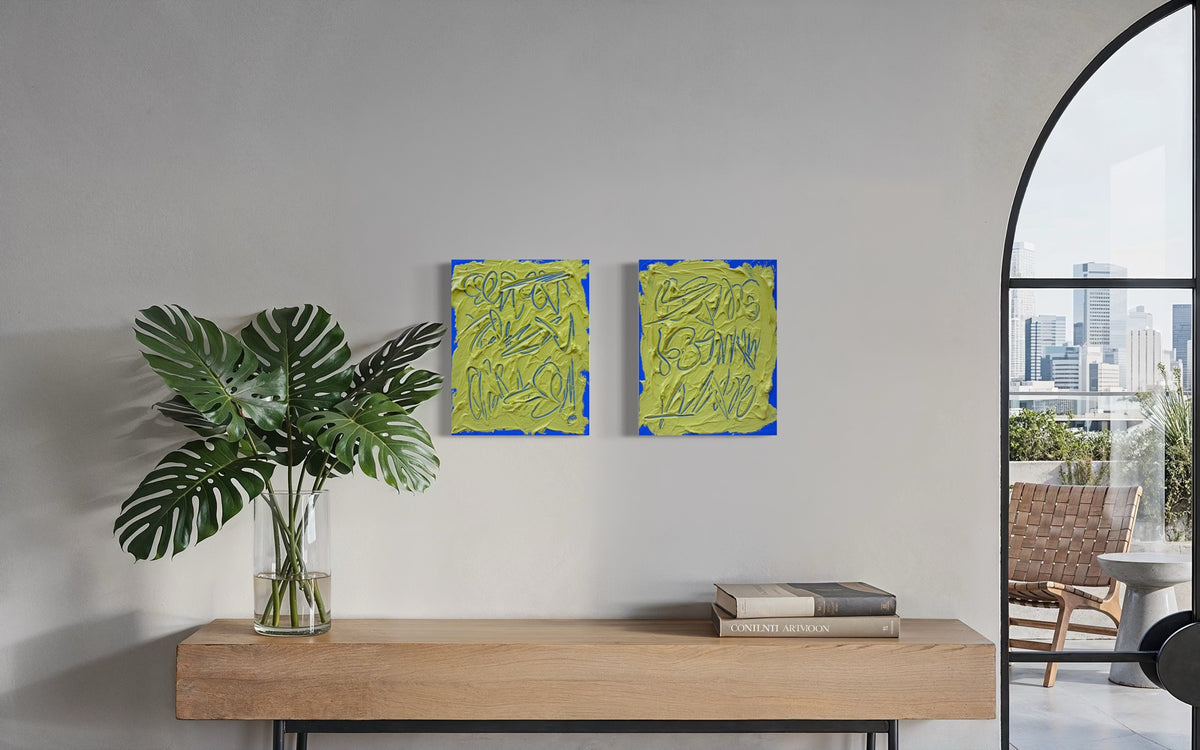 Two abstract text based painting featuring a thick, yellow-green impasto texture with blue abstract scribbled lines hang on a light gray wall above a wooden console table with a potted plant and books. A large arched window on the right shows a city view and outdoor chair.