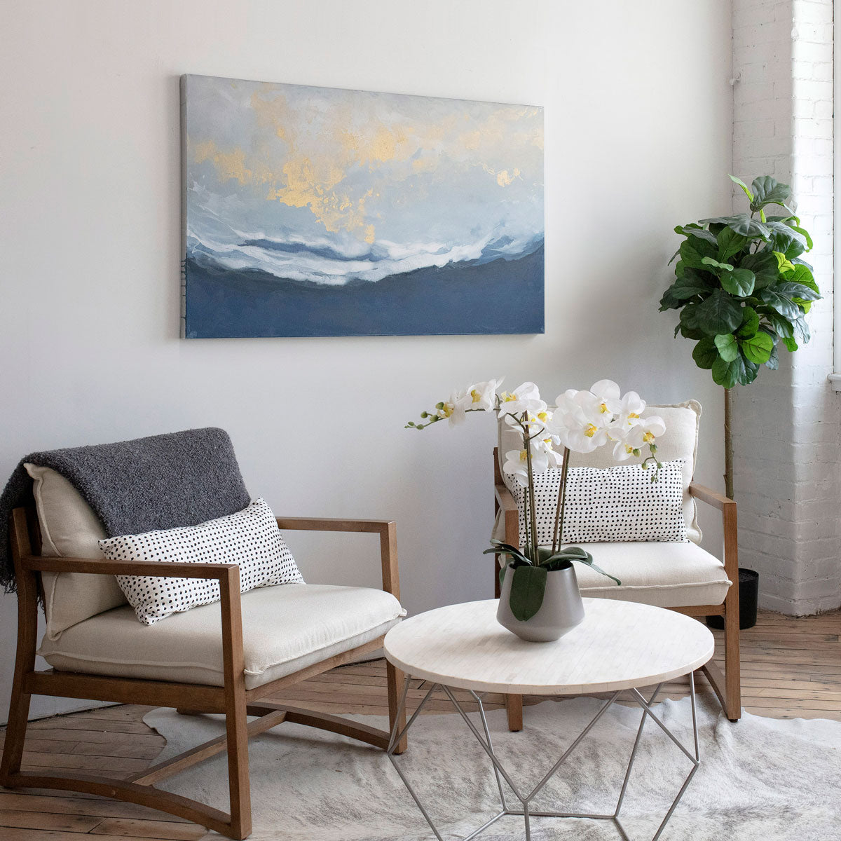 An abstract blue, white, and gold painting hangs on a light wall above modern armchairs and a coffee table with potted orchid. 