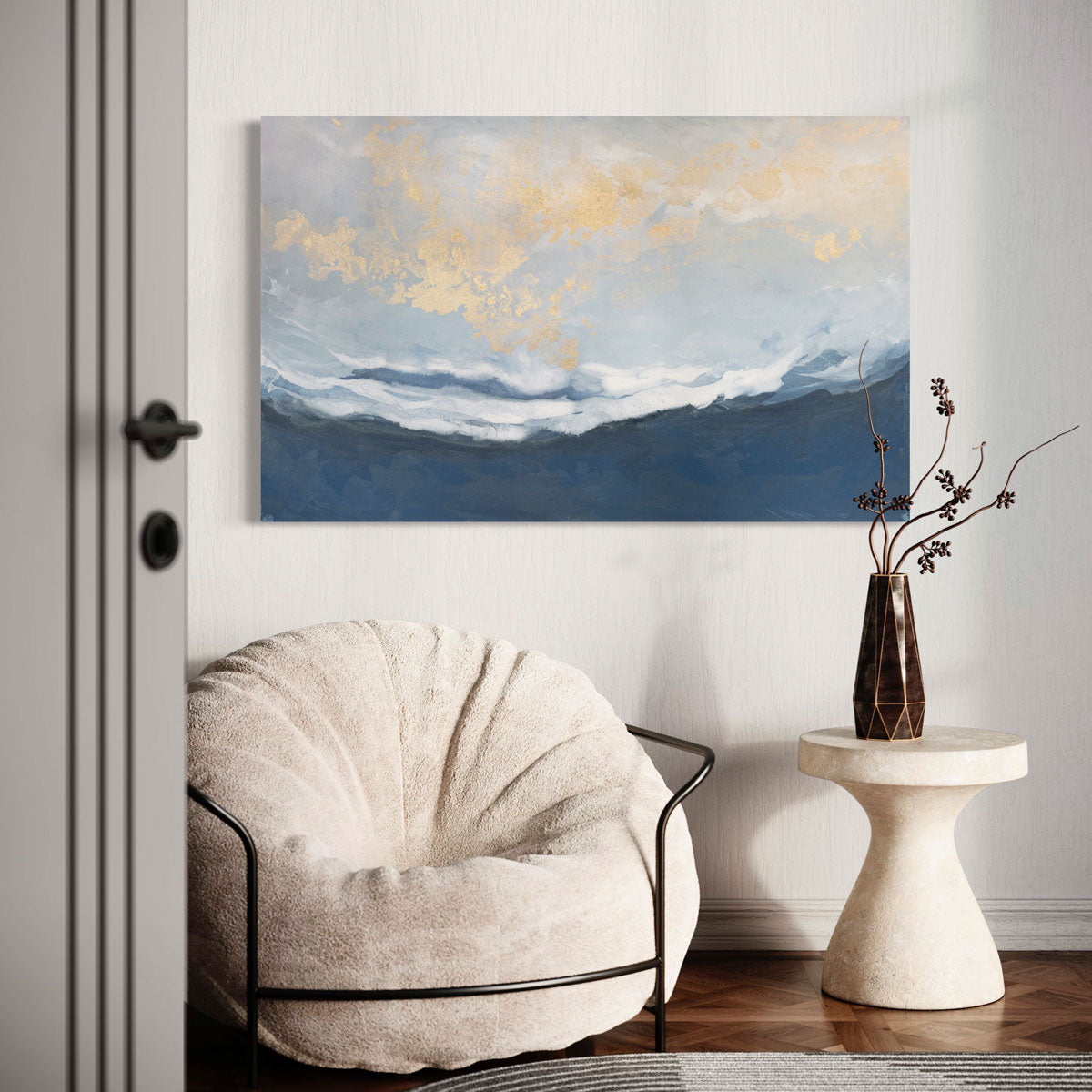 An abstract blue, white, and gold painting hangs on a light wall above a beige contemporary stuffed chair and modern end table.