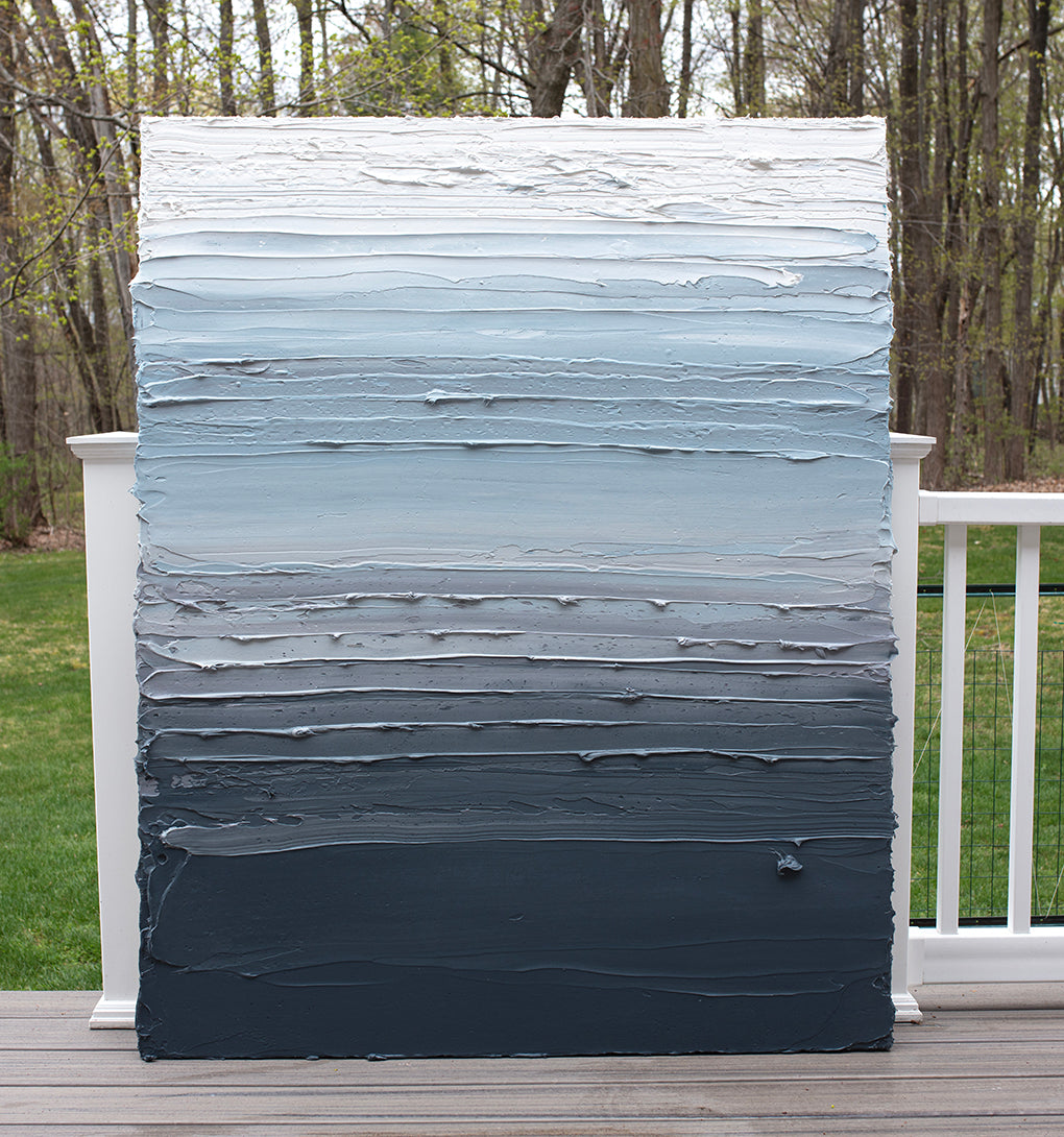 A dark grey blue, light blue, blue green, and white thickly textured abstract painting leaning on a deck outside in natural light by Teodora Guererra.