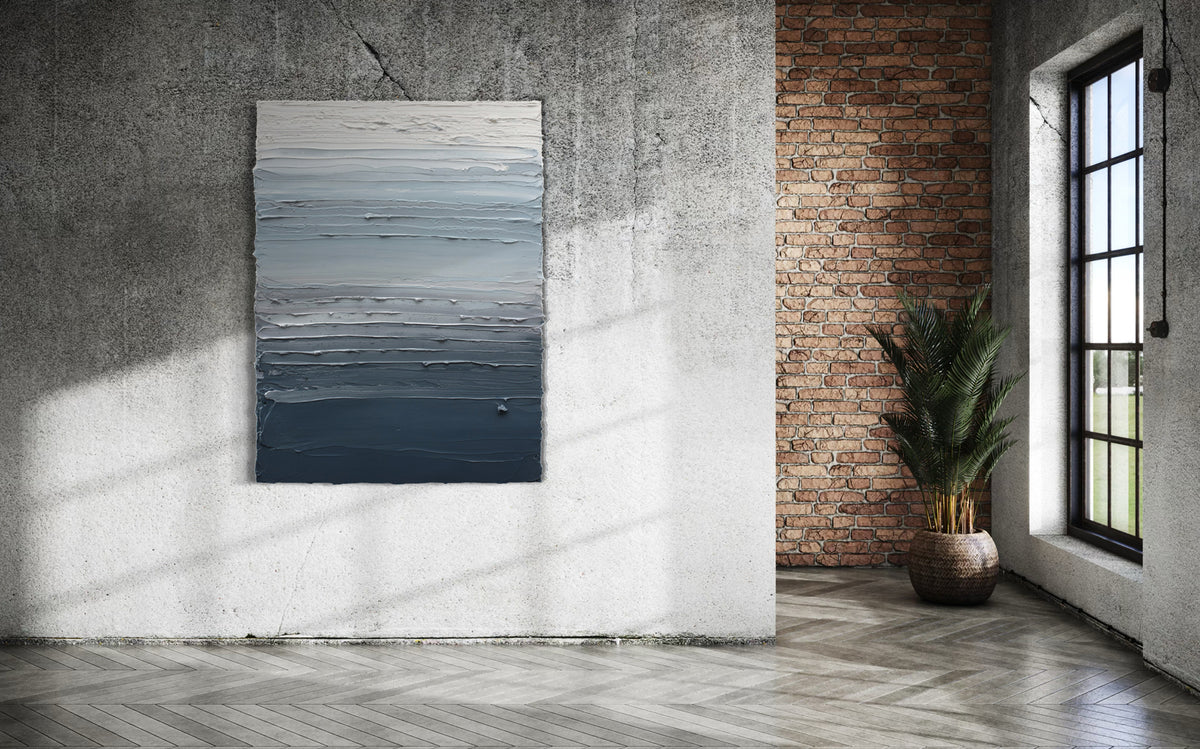 A dark grey blue, light blue, blue green, and white thickly textured abstract painting hanging on a cement wall with a window and potted plant to the right.