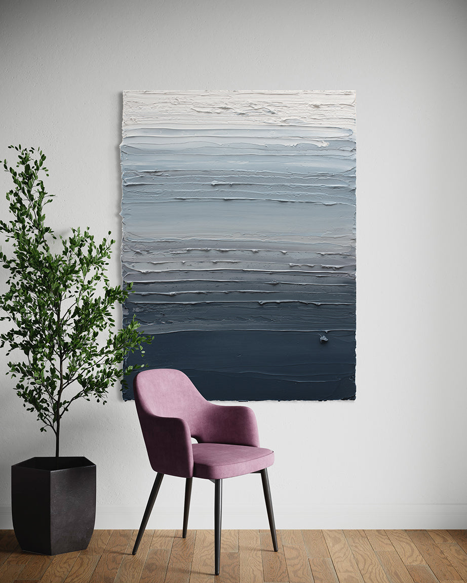 A dark grey blue, light blue, blue green, and white thickly textured abstract painting hangs on a white wall by Teodora Guererra. In front of the painting is a chair with dark metal legs covered in a pink/lavender colored fabric. To the left of the chair is a potted fica tree.