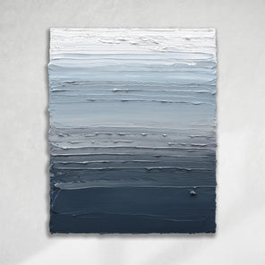 A vertical thickly textured abstract painting in bands of dark blue grey, light blue, blue green, and white hanging on a white wall in natural light by artist Teodora Guererra.