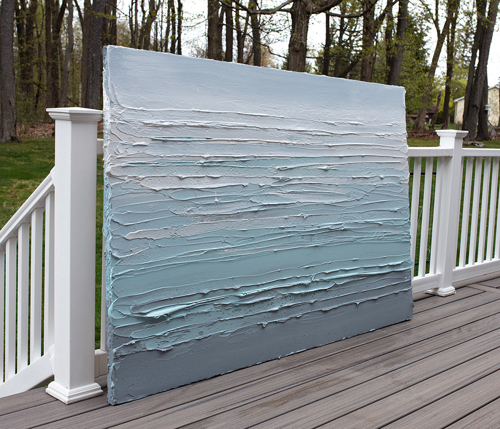 An abstract painting with turquoise impasto brushstrokes in a color gradient is sitting on a deck on an angle in the artists studio.