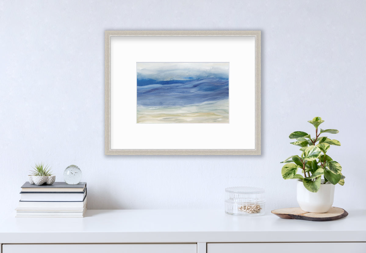A blue, white and beige abstract print in a silver frame with a mat hangs on a white wall above a white surface with a stack of books, a round clear container with gold accessories and a potted plant.