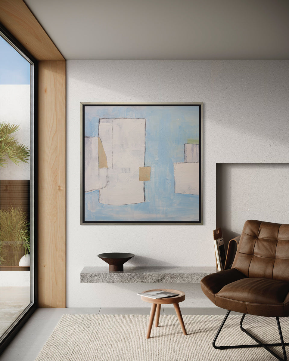 A blue, white and beige abstract print in a silver floater frame hangs on a white wall above a granite floating shelf, a small side table and a brown leather chair.