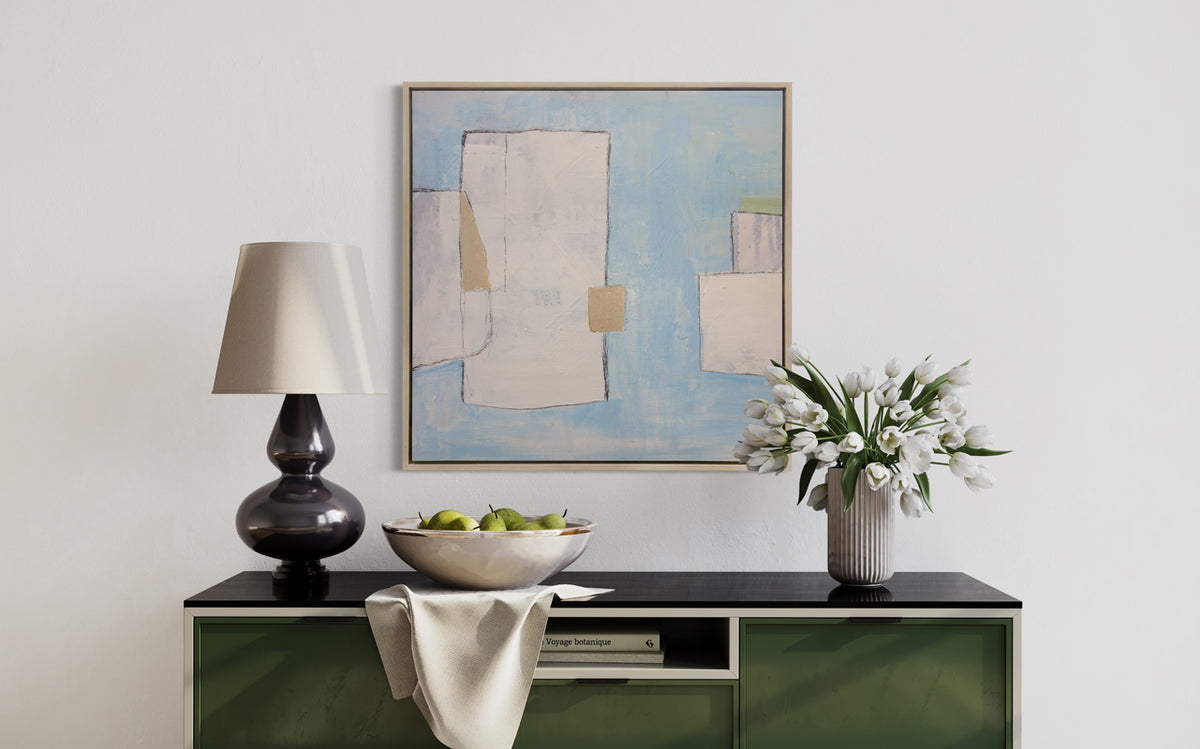 A blue, white and beige abstract print in a white oak wood floater frame hangs on a white wall above a green and black credenza with a black table lamp, a bowl of pears and a white vase with white tulips.