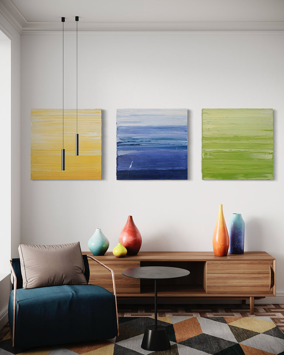 Three textured abstract paintings, one in yellow and white, another in blue and white and the last in chartreuse and white by Teodora Guererra hang on a wall over a credenza with bright colored ceramic pots.